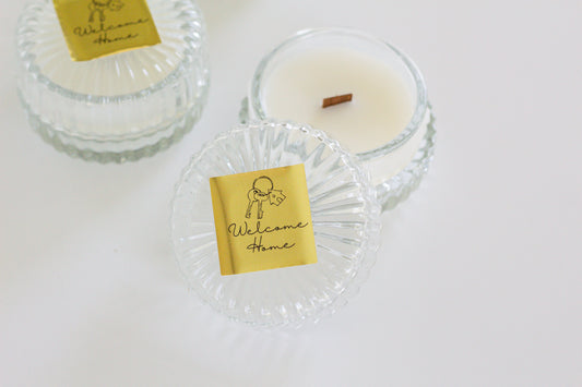 Two clear glass jar candles with gold 'Welcome Home' labels are arranged on a white surface. One candle is open, showing white wax and a small brown wood wick, while the other one is closed.