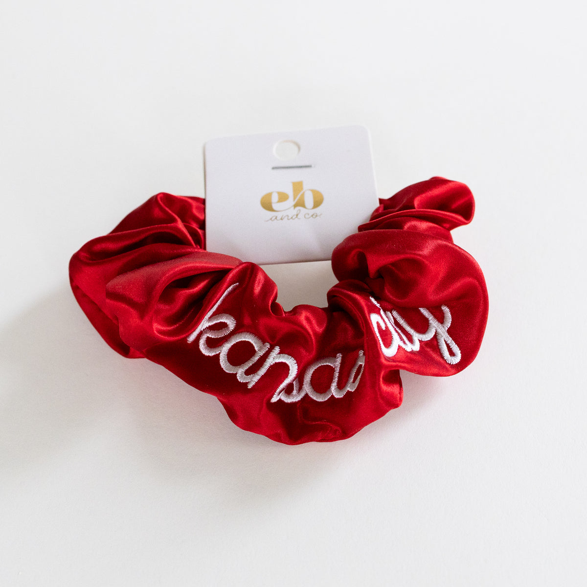 A red satin hair scrunchie with white embroidered cursive text reading 'kansas city', attached to a white cardboard tag with a gold logo, photographed on a clean white background.