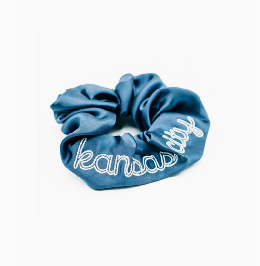 A blue satin scrunchie with 'Kansas City' embroidered in white script across the front, displayed on a clean white background.
