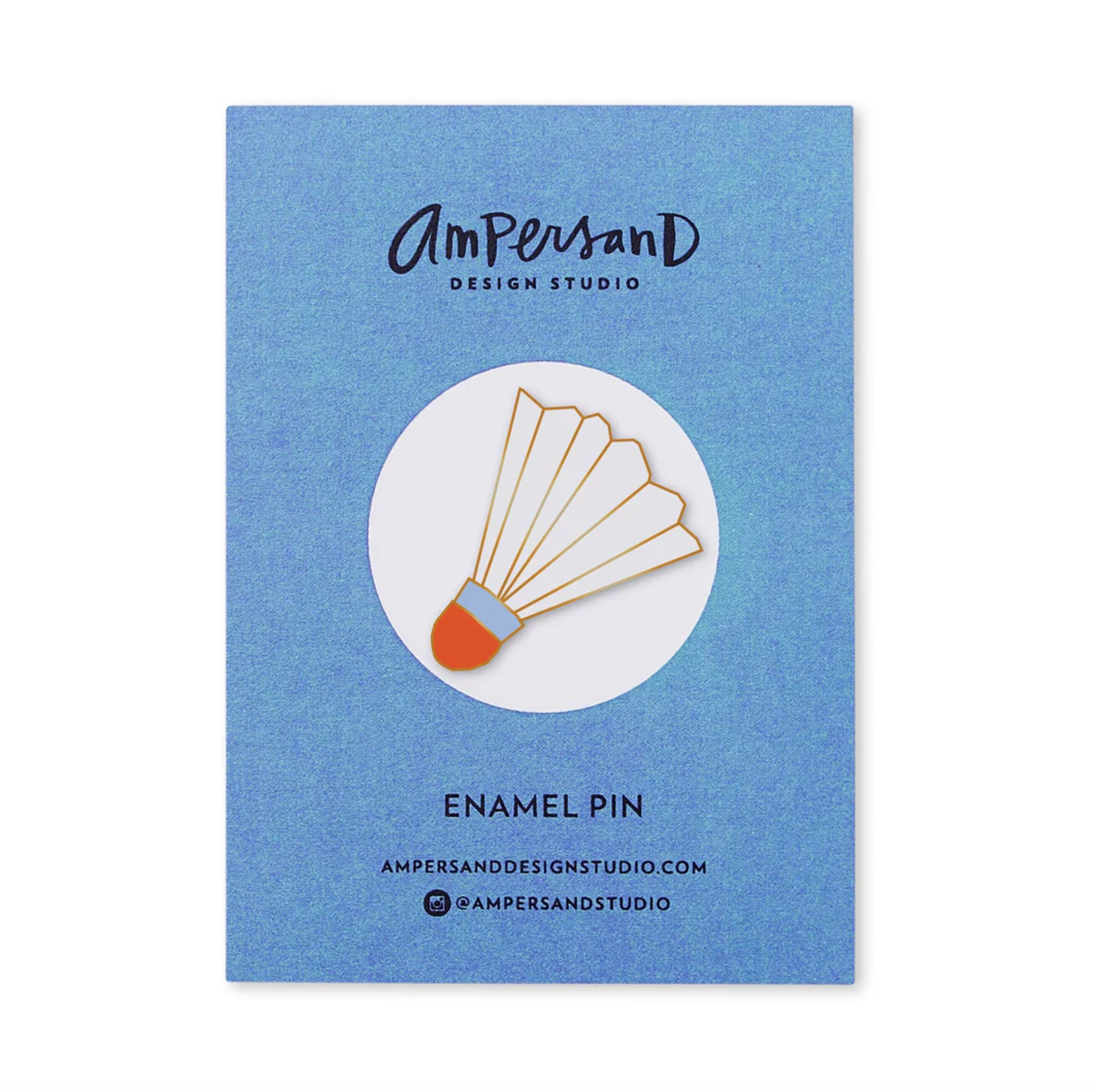 An Ampersand Design Studio enamel pin of a shuttlecock, shown in its blue textured backing card packaging. The pin features a gold-tone metal shuttlecock body with a white feather section, a light blue band, and an orange-brown head.