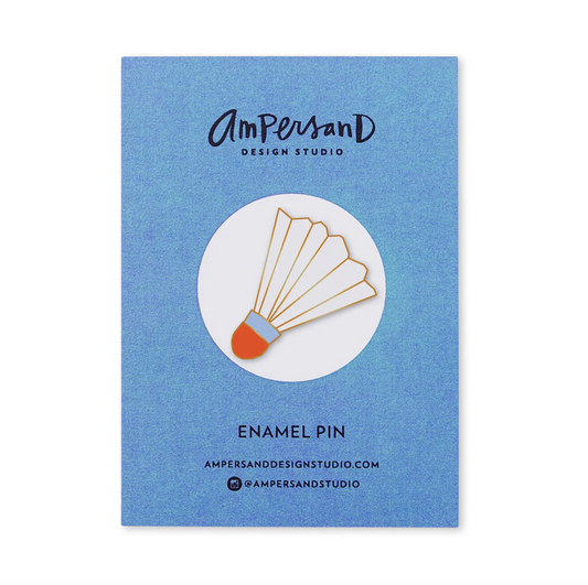 An Ampersand Design Studio enamel pin of a shuttlecock, shown in its blue textured backing card packaging. The pin features a gold-tone metal shuttlecock body with a white feather section, a light blue band, and an orange-brown head.