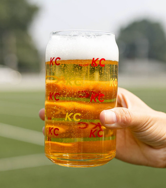 A hand holding a clear glass can-shaped mug filled with golden beer and topped with white foam. The mug has red 'KC' logos printed on it, and the background is a blurred outdoor field.