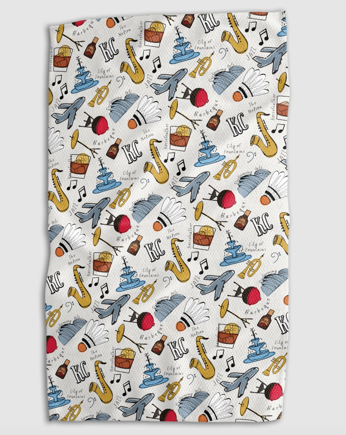 A rectangular kitchen towel with a white background and a colorful repeating pattern of Kansas City monuments, cocktail glasses, saxophones, BBQ sauce bottles, and the text 'KC'.