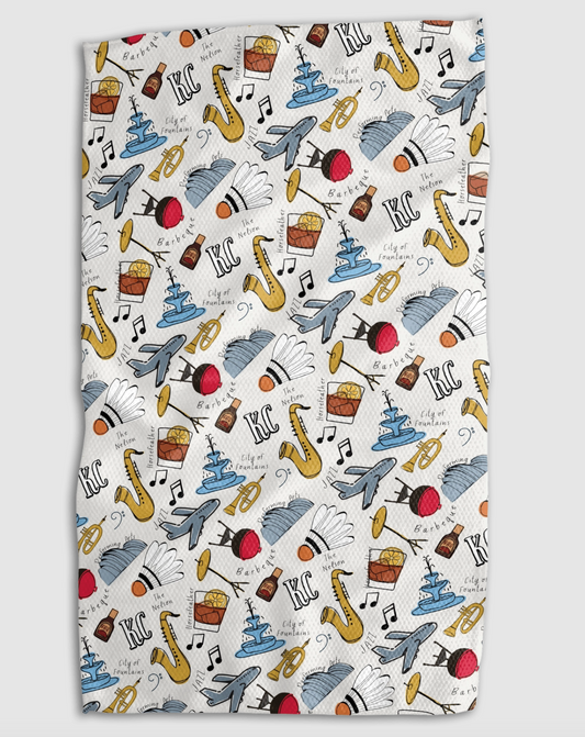 A rectangular kitchen towel with a white background and a colorful repeating pattern of Kansas City monuments, cocktail glasses, saxophones, BBQ sauce bottles, and the text 'KC'.