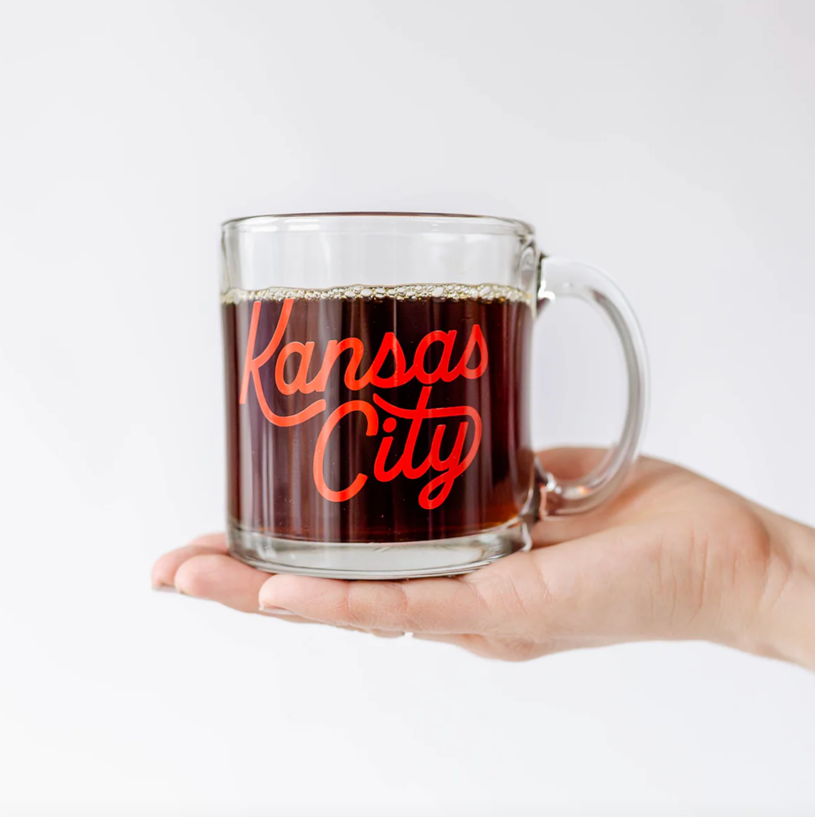 A hand holding a clear glass mug filled with dark coffee, featuring the words 'Kansas City' in red script on the front, against a plain white background.