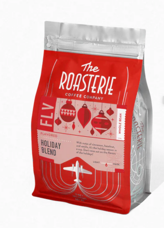 A stand-up coffee bag from The Roasterie Coffee Company, featuring a holiday-themed design with red and white graphics, Christmas ornaments, and an airplane illustration on a white background.