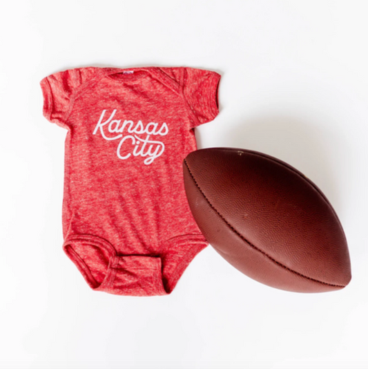 A heather red short-sleeve baby bodysuit with 'Kansas City' printed in white script, displayed next to a brown leather football on a clean white background.