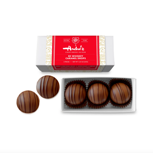 An overhead shot of Andre's Confiserie Suisse KC Whiskey Caramel Drops with a 3 piece box and 2 pieces sitting outside of the box on a white background. Each whiskey caramel drop is a round chocolate piece with chocolate drizzle on it. 