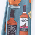 Load image into Gallery viewer, A folded towel, featuring a blue-green background with a pattern of illustrated whiskey bottles and labels in orange, black, white, and yellow.
