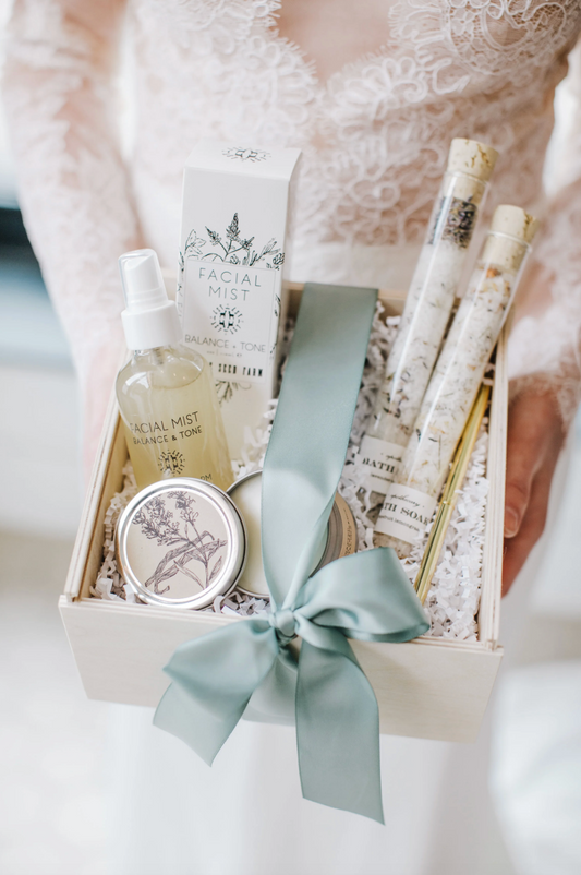 A self-care-inspired gift set containing facial mist, a candle, and tubes of bath soak salts, presented in a cream-colored box with a sage green ribbon, held by a person in a white lace wedding dress.