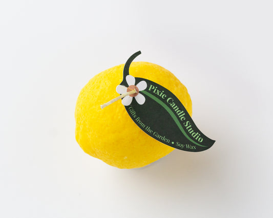A bright yellow lemon-shaped candle with a green petal tag that reads Pixie Candle Studio. 