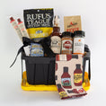 Load image into Gallery viewer, A barbecue gift basket in a black and yellow utility tote, a bag of Rufus Teague Spittin' Seeds sunflowers, a bottle of BBQ Sauce, a jars of spices and seasonings, and a patterned towel with barbecue bottle illustrations, photographed on a white background.
