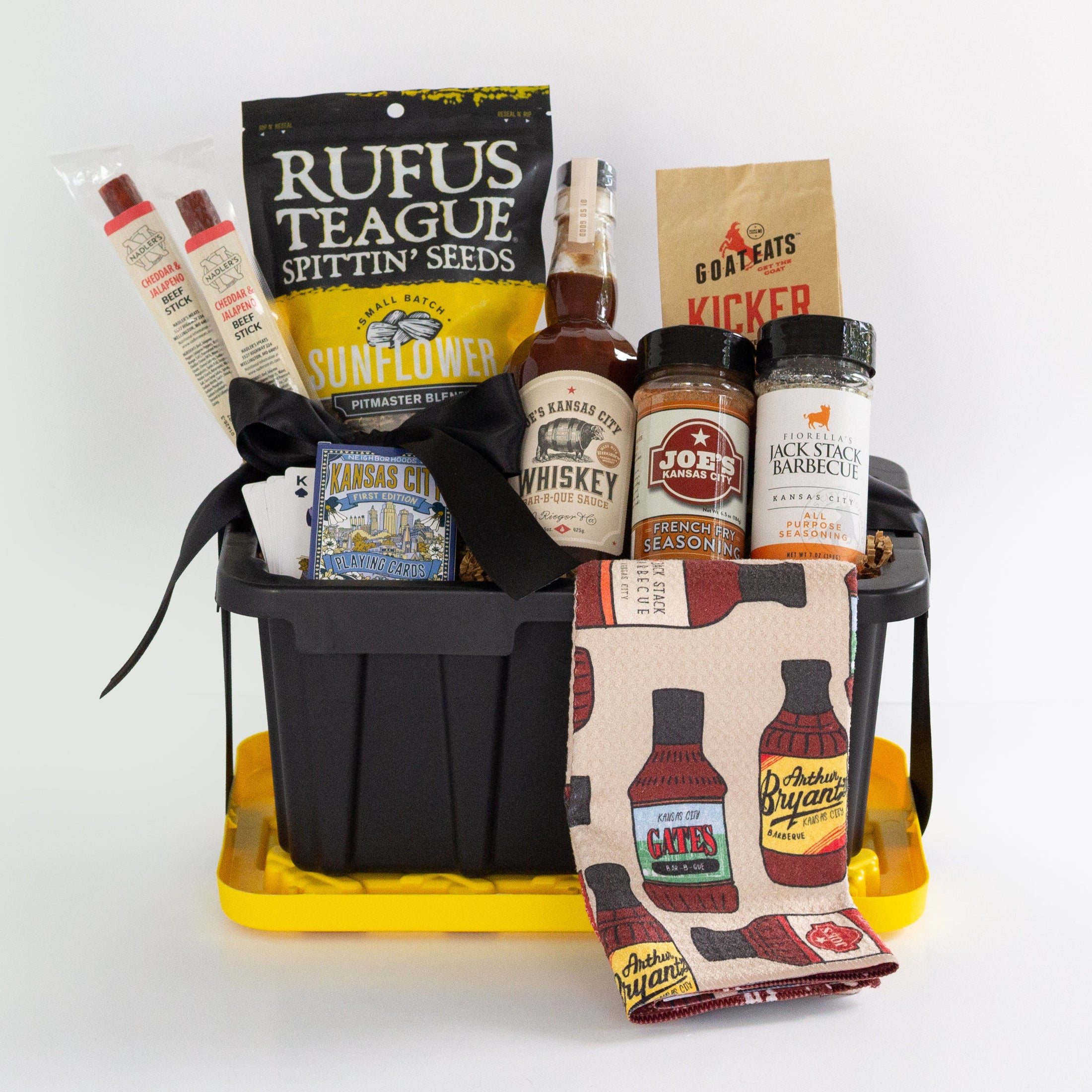 A barbecue gift basket in a black and yellow utility tote, a bag of Rufus Teague Spittin' Seeds sunflowers, a bottle of BBQ Sauce, a jars of spices and seasonings, and a patterned towel with barbecue bottle illustrations, photographed on a white background.