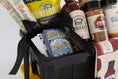 Load image into Gallery viewer, A barbecue gift basket in a black and yellow utility tote, a bag of Rufus Teague Spittin' Seeds sunflowers, a bottle of BBQ Sauce, a jars of spices and seasonings, and a patterned towel with barbecue bottle illustrations, photographed on a white background.
