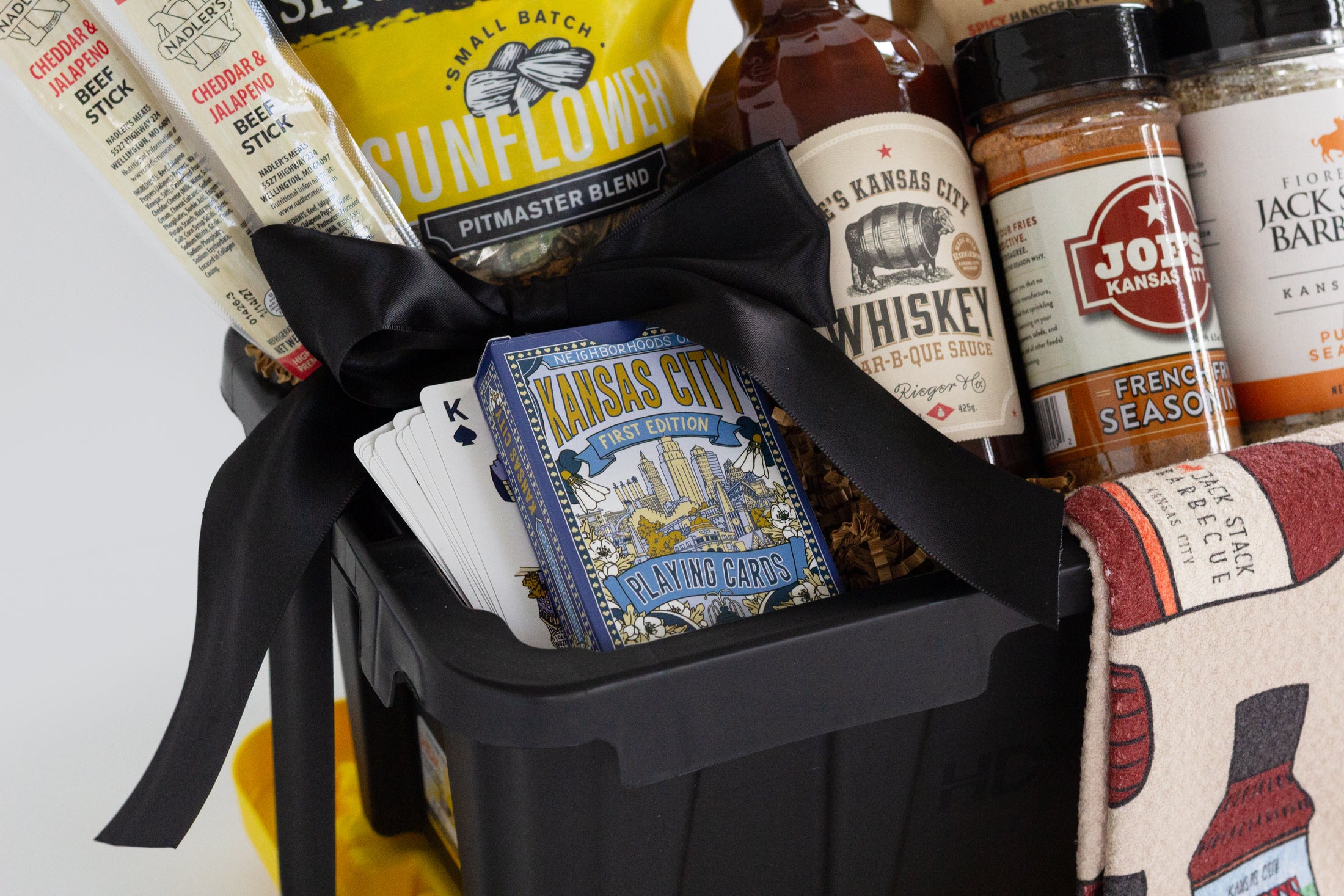 A barbecue gift basket in a black and yellow utility tote, a bag of Rufus Teague Spittin' Seeds sunflowers, a bottle of BBQ Sauce, a jars of spices and seasonings, and a patterned towel with barbecue bottle illustrations, photographed on a white background.