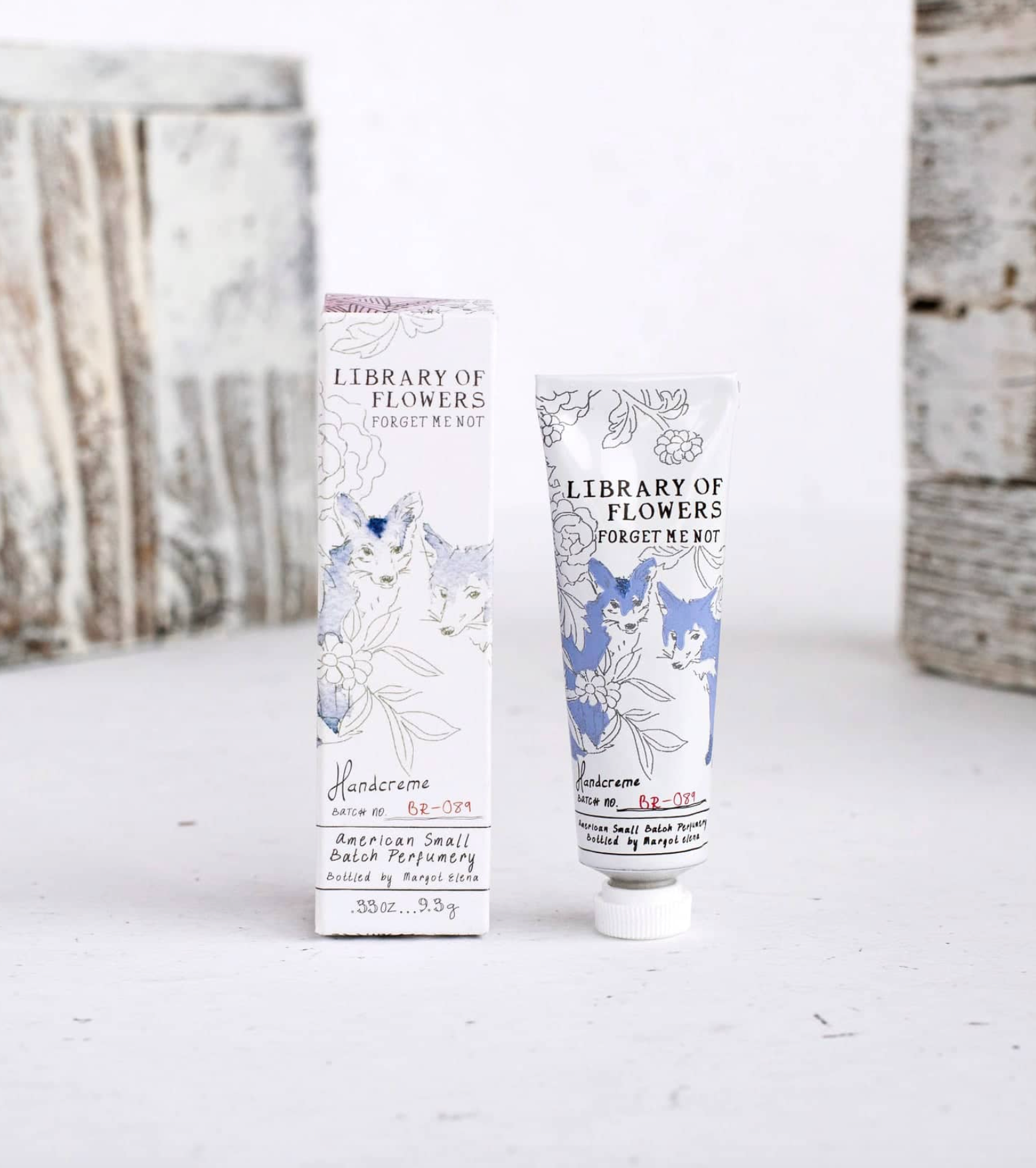 A tube and matching box of Library of Flowers Forget Me Not hand cream, featuring white packaging with blue and black botanical illustrations, set against a rustic white wooden background.