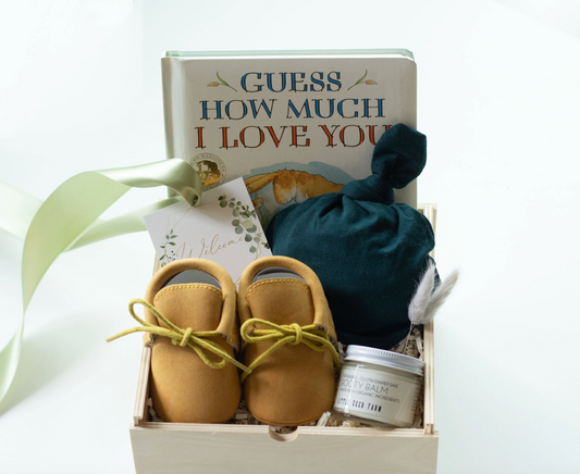 A baby gift set arranged in a light wood box on a white background. The set includes a book titled 'GUESS HOW MUCH I LOVE YOU', a pair of tan suede moccasins, a dark teal fabric knit cap, and a small white jar of Booty Balm.