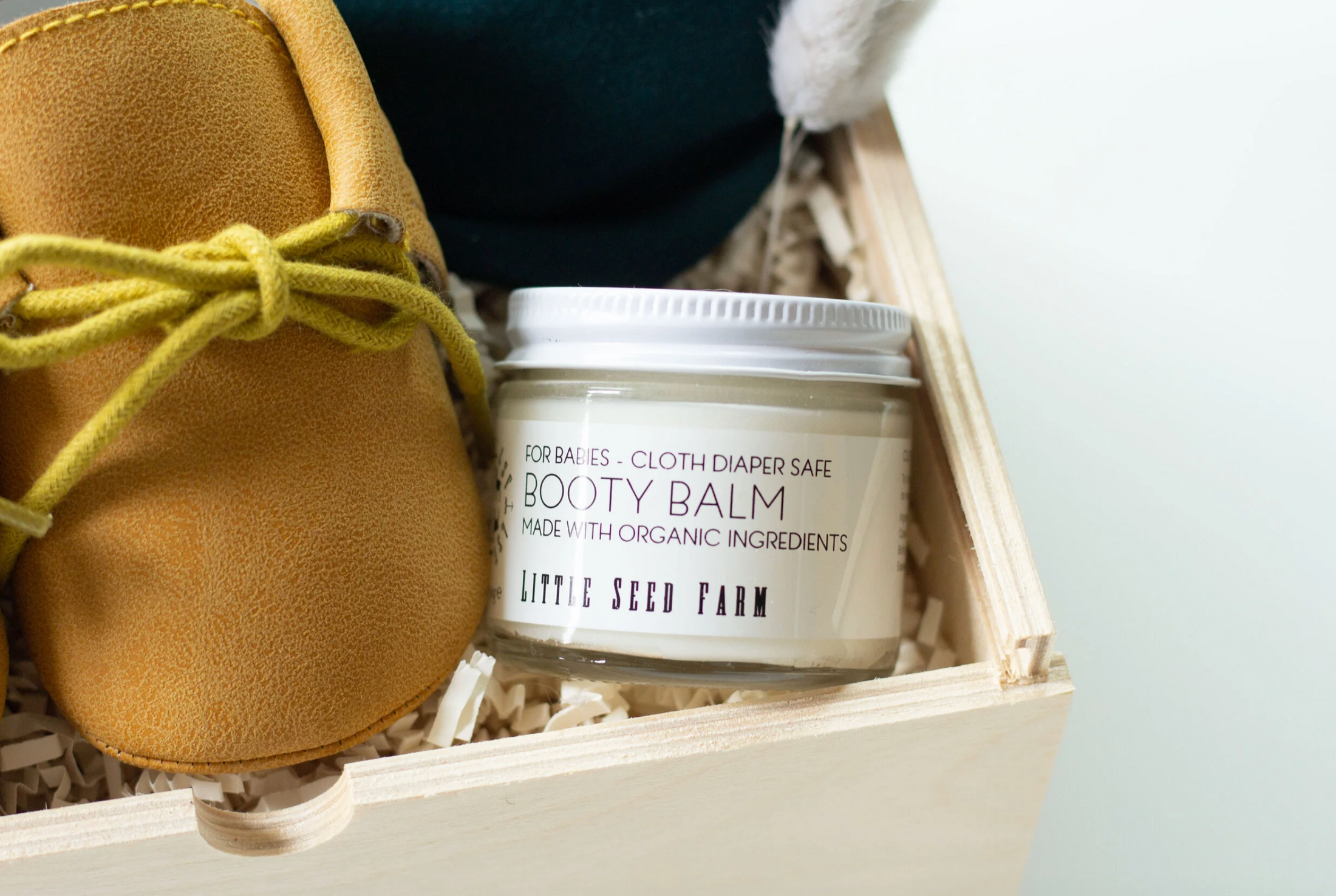 A jar of Little Seed Farm Booty Balm placed in a wooden gift box with shredded paper, next to a tan suede bootie with a yellow-green laces, against a light gray background.