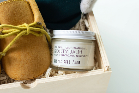 A jar of Little Seed Farm Booty Balm placed in a wooden gift box with shredded paper, next to a tan suede bootie with a yellow-green laces, against a light gray background.