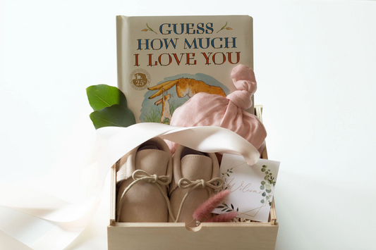 A baby shower gift box containing a book titled 'GUESS HOW MUCH I LOVE YOU', a pair of beige baby booties, a light pink knotted knit cap, a small card, and green leaves, set against a clean white background.