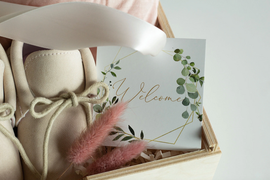 A close-up of a gift box containing cream-colored baby booties tied with beige rope, a white welcome card with green botanical illustrations and gold script, and pink dried grass stems on a light background.