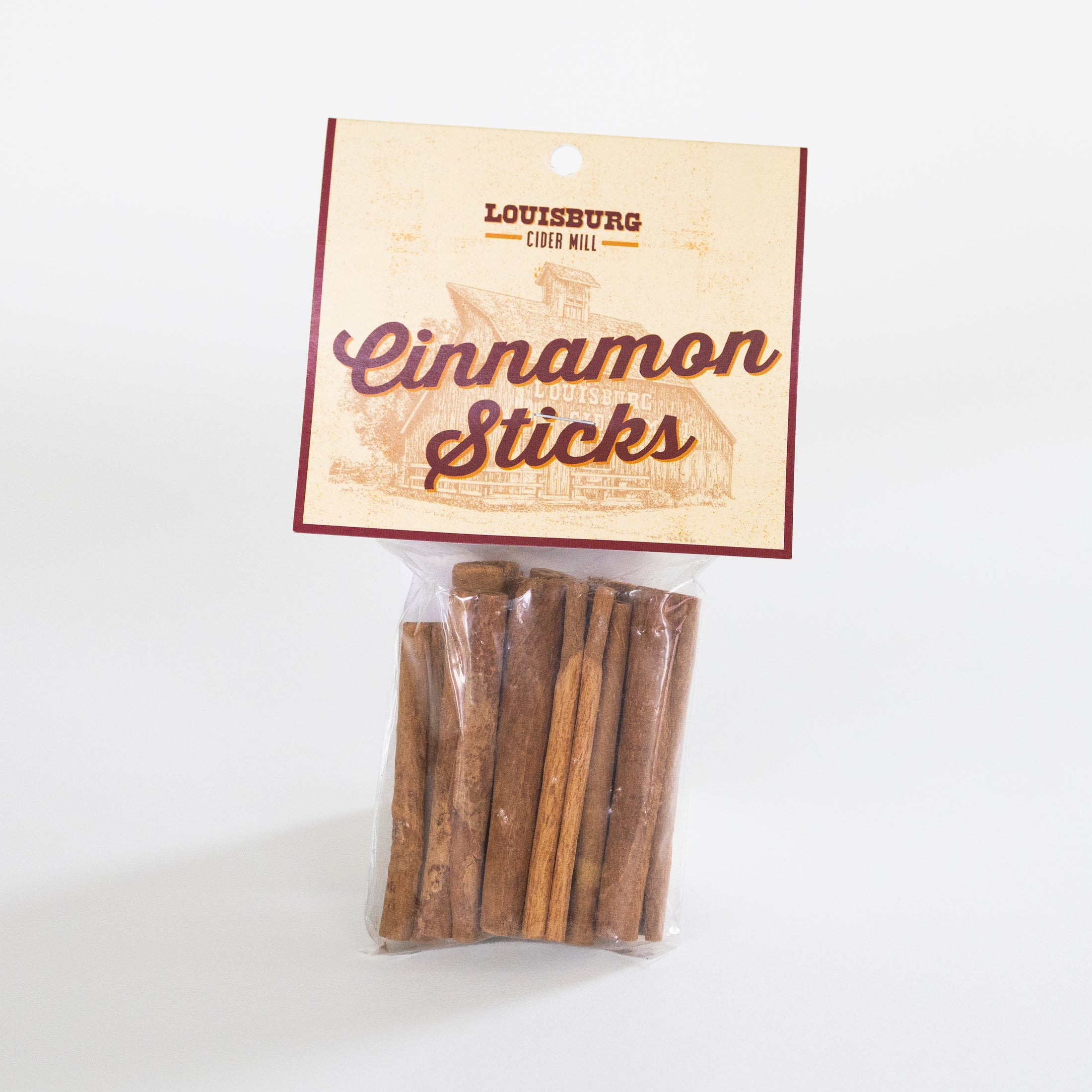 A clear plastic bag containing several cinnamon sticks, attached to a cream-colored paper header card with the text 'Louisburg Cider Mill Cinnamon Sticks' and a sepia illustration of the Louisburg Cider Mill.