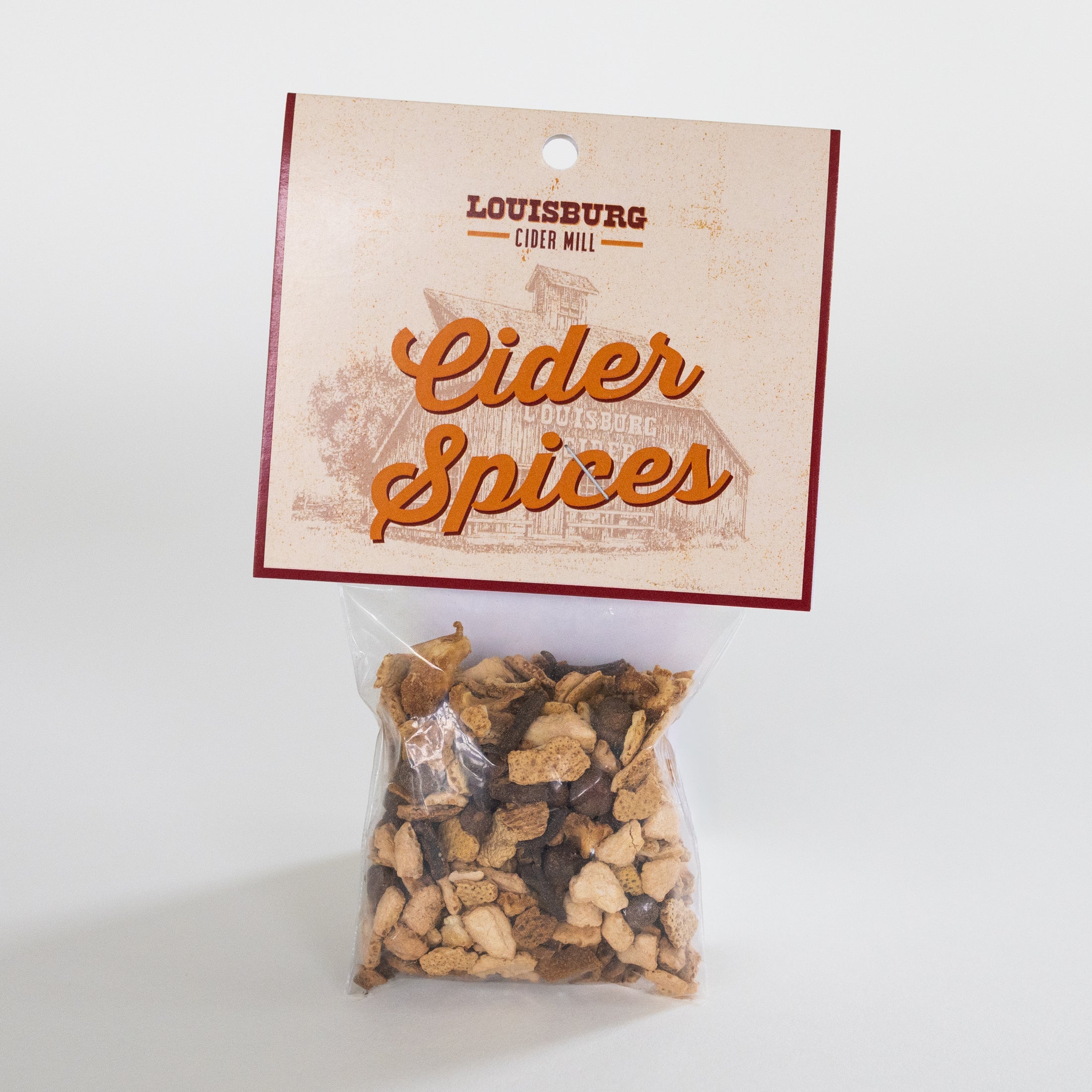 A clear plastic bag filled with loose-leaf cider spices, attached to a printed kraft paper header card that reads 'LOUISBURG CIDER MILL' and 'Cider Spices' in orange script, photographed on a light gray background.