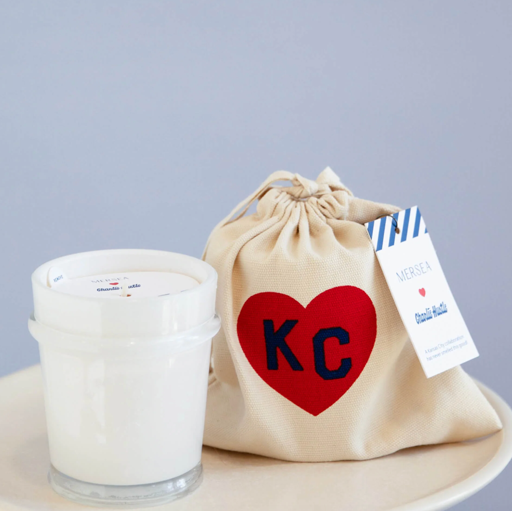 A white candle in a clear glass jar sits beside a beige drawstring gift bag with a red heart and navy letters 'KC' on the front. 