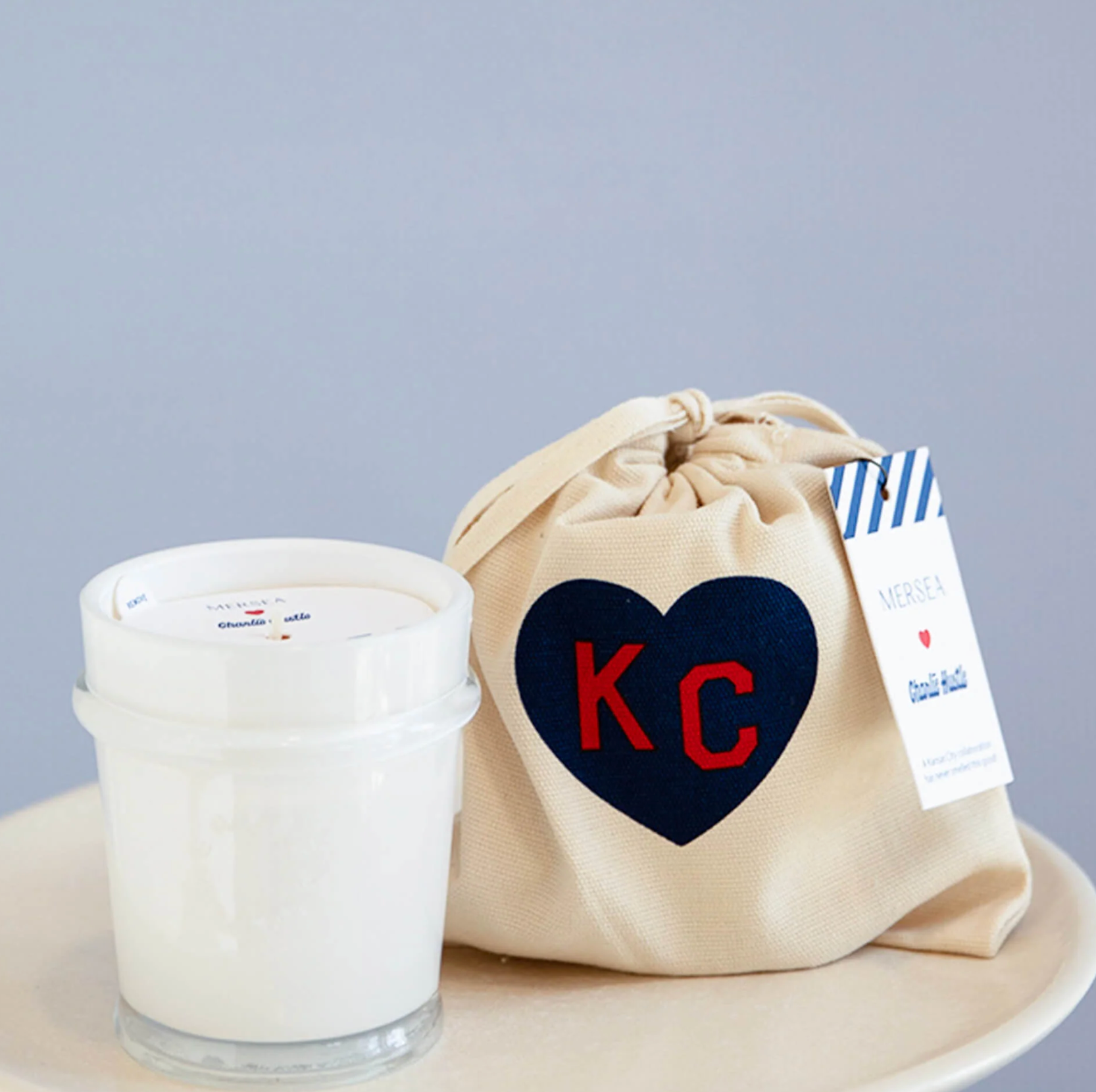 A white candle jar next to a beige fabric drawstring bag with a navy heart patch featuring red letters 'KC' and a white tag, set on a light surface against a pale blue-gray background.