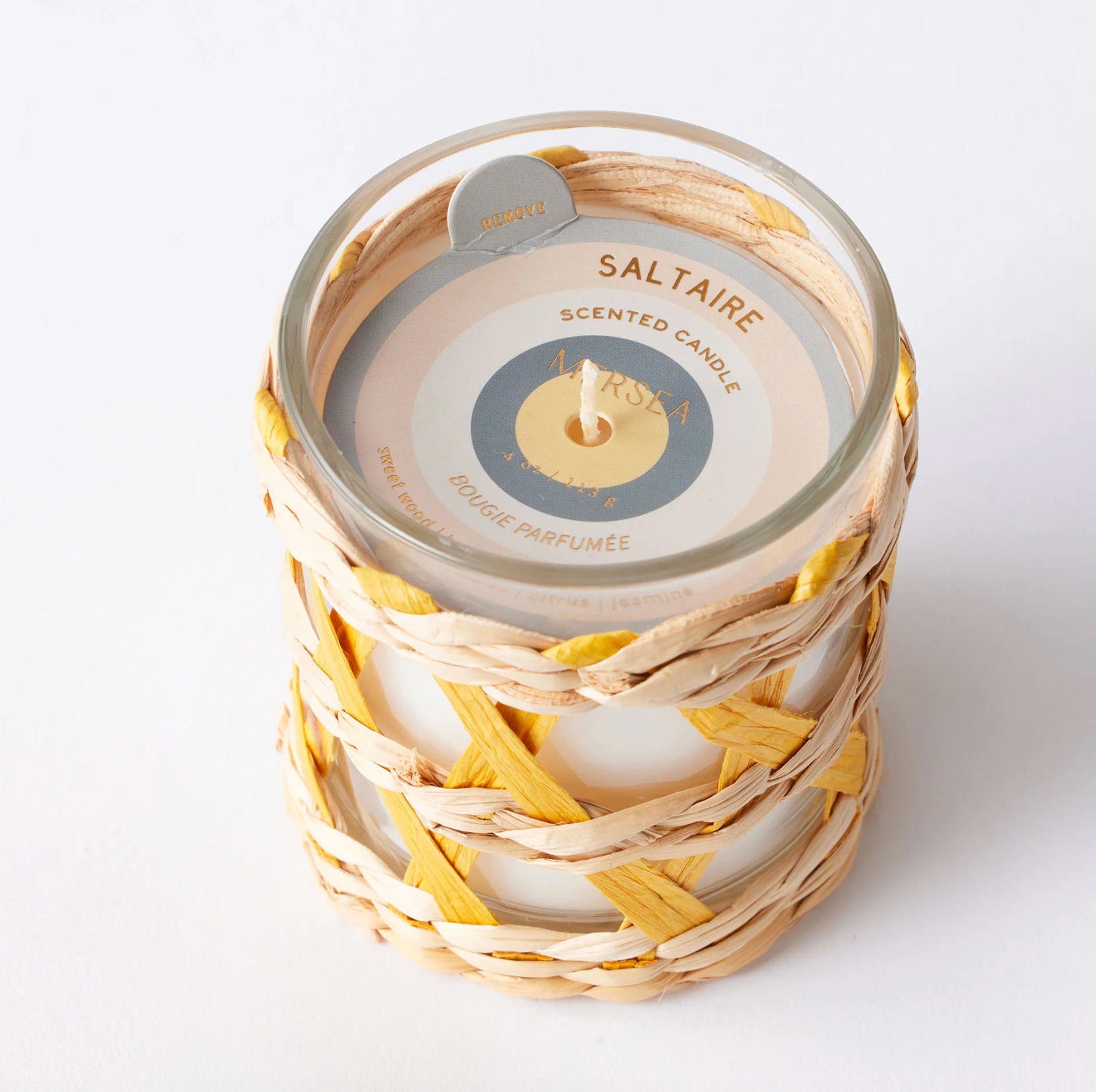 A MERSEA co. Saltaire scented candle in a clear glass jar wrapped in a natural woven straw basket, on a clean white background.