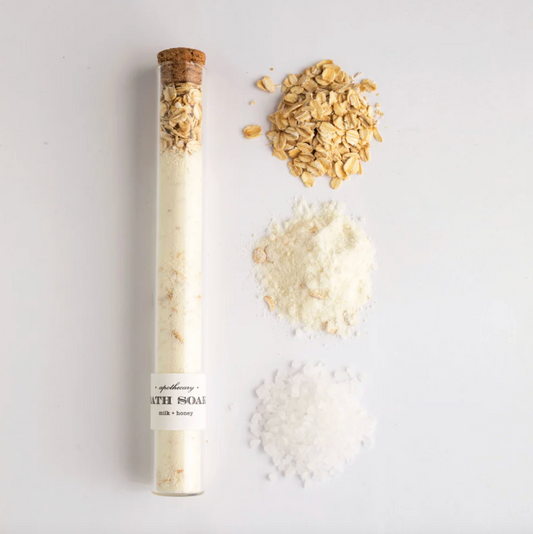 A tube of bath soak vial on a white background next to the bath soak contents which are oatmeal, milk powder, and bath salts. 