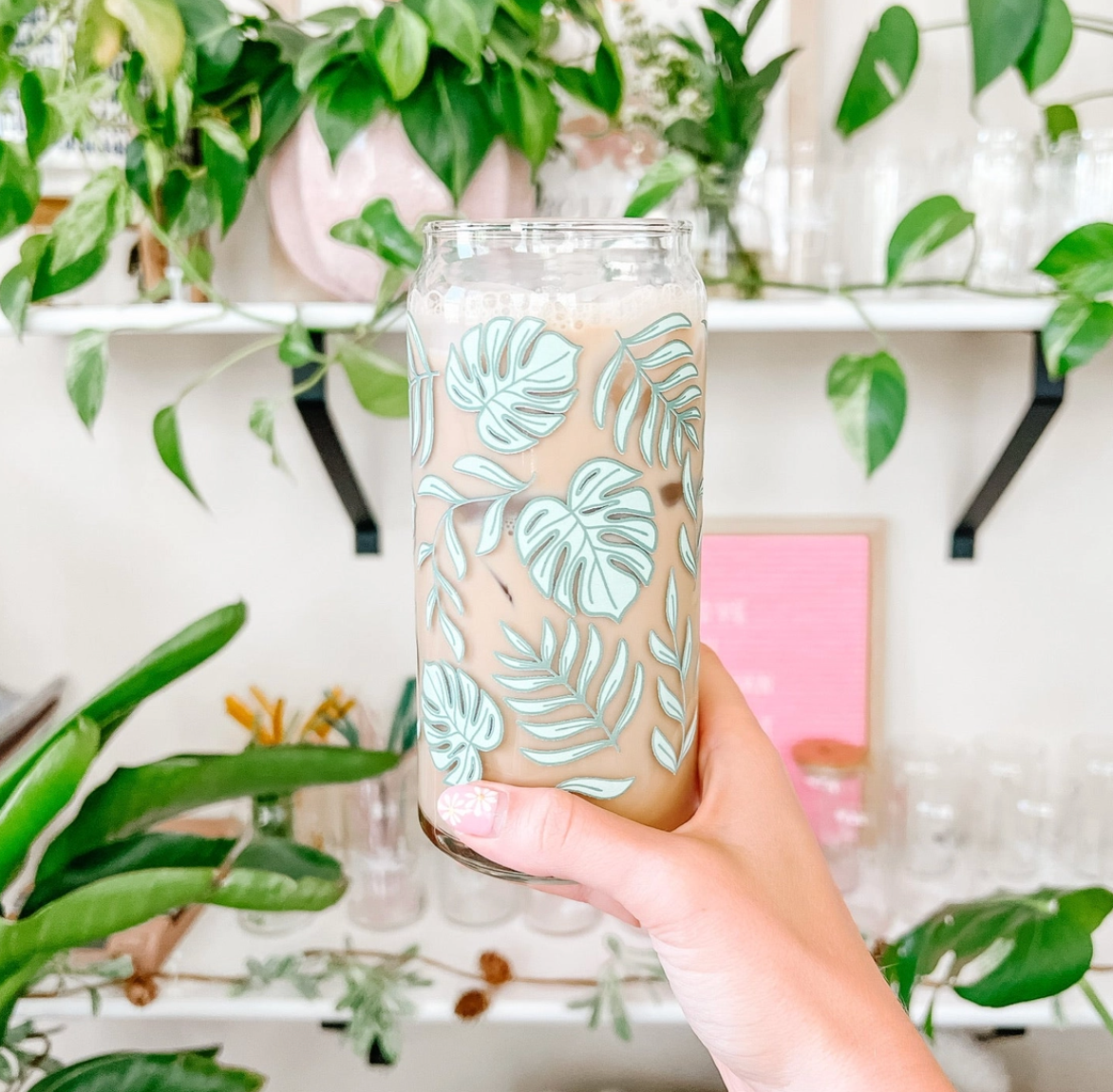 A hand holds a clear glass can-shaped tumbler decorated with a light green monstera leaf pattern, filled with an iced coffee drink, set against a background of green houseplants on white shelves.