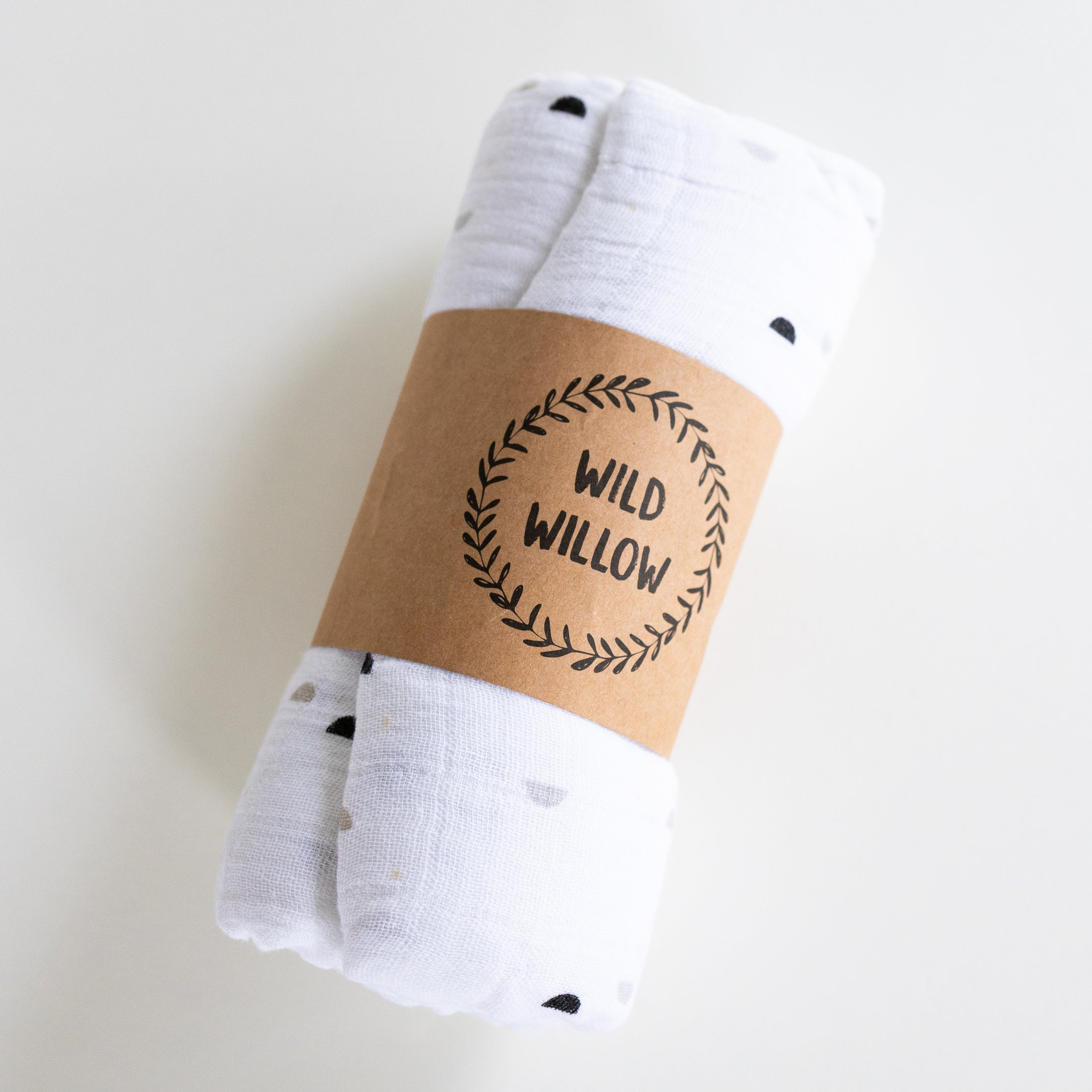 A rolled white muslin baby swaddle blanket with small black and gray polka dots, wrapped in a brown kraft paper band printed with the brand name 'WILD WILLOW'.