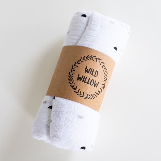 A rolled white muslin baby swaddle blanket with small black and gray polka dots, wrapped in a brown kraft paper band printed with the brand name 'WILD WILLOW'.