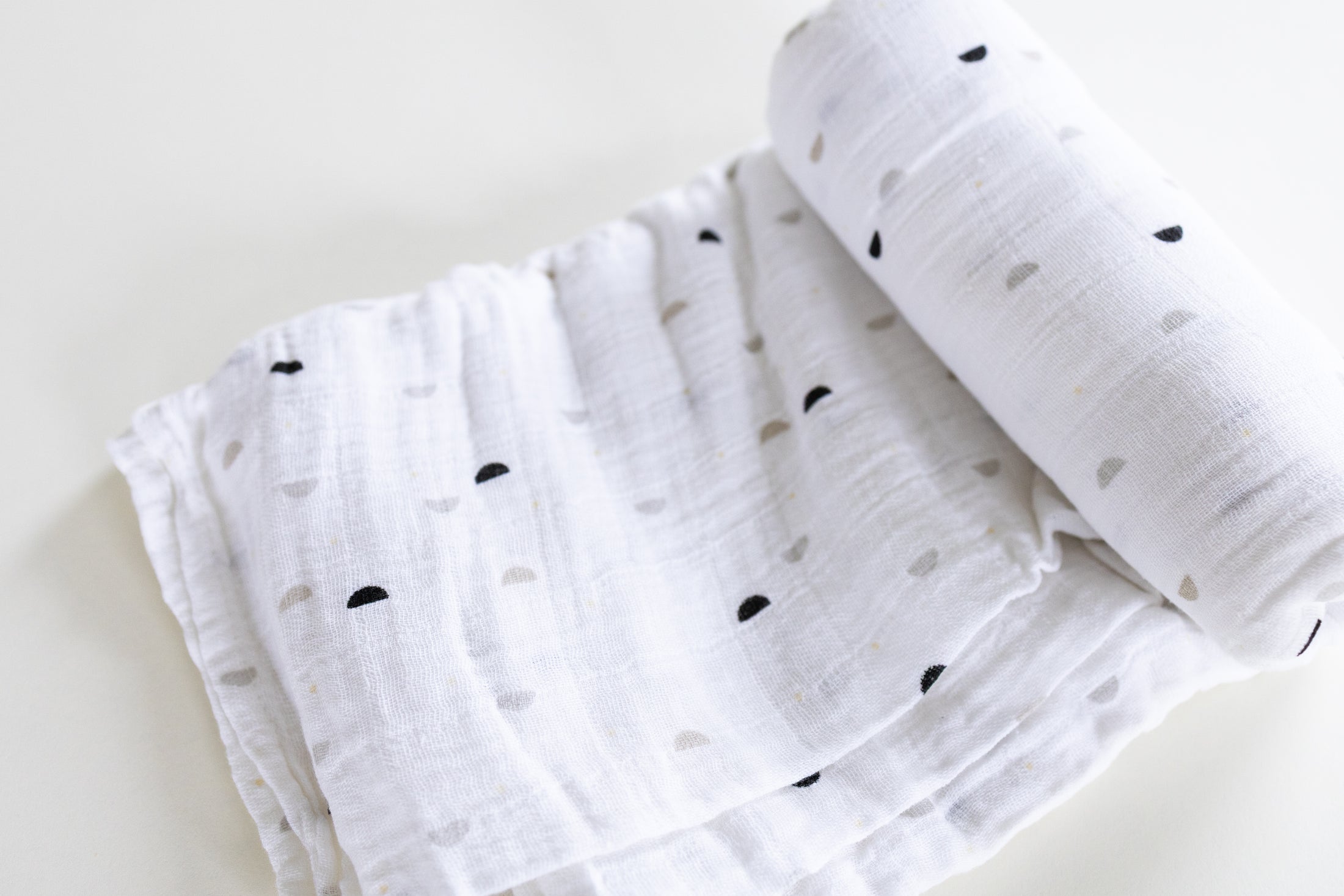 A white muslin blanket with a small black and gray heart pattern, folded and rolled on a light cream background.