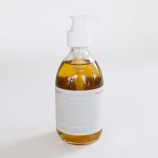 A clear glass pump bottle filled with amber-colored hand soap, featuring a white pump dispenser and a white label with fine blue and red text, set against a plain white background.