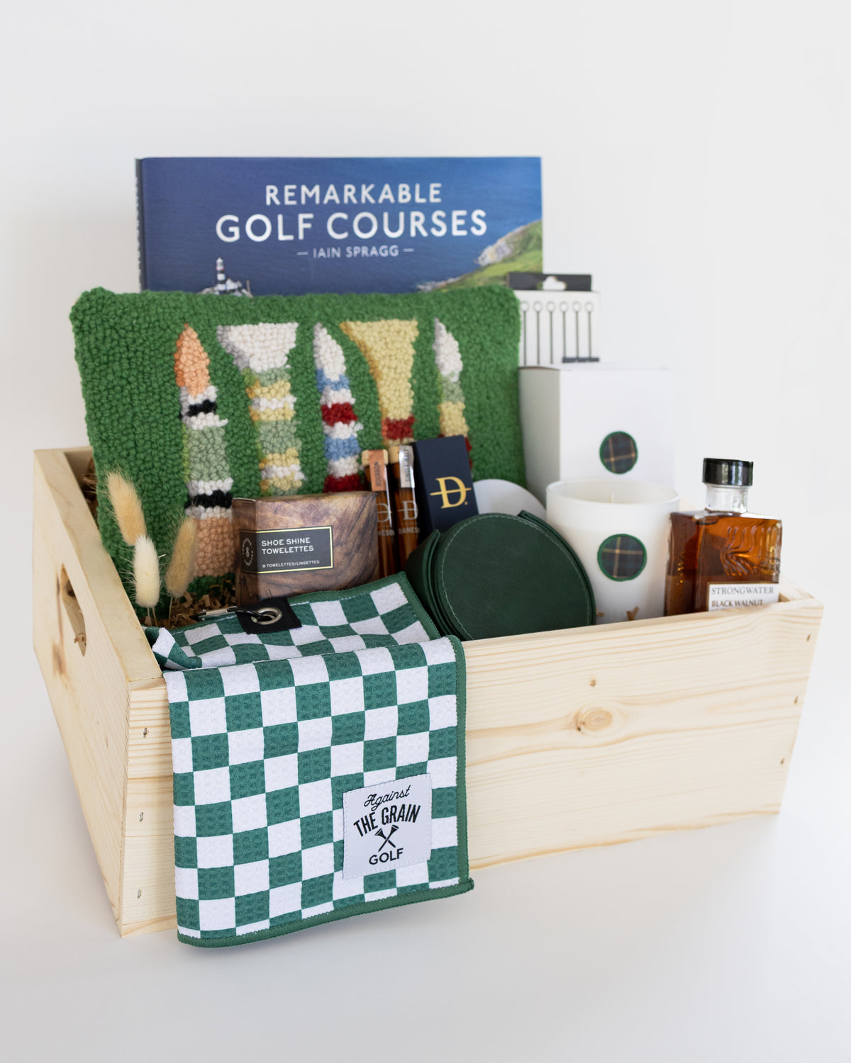 A wooden gift basket filled with golf-themed items, including a book titled 'Remarkable Golf Courses' by Iain Spragg, a green textured pillow with multi-colored golf tees, a green checkered towels, a white candle with a green plaid wax se