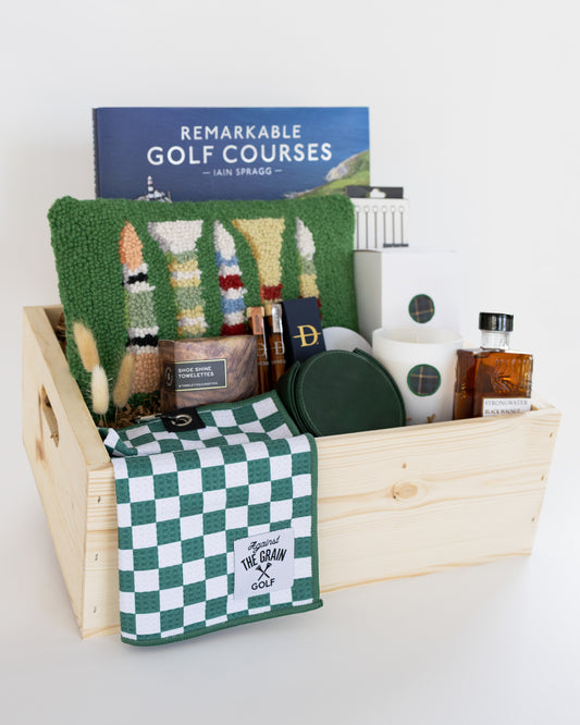 A wooden gift basket filled with golf-themed items, including a book titled 'Remarkable Golf Courses' by Iain Spragg, a green textured pillow with multi-colored golf tees, a green checkered towels, a white candle with a green plaid wax se