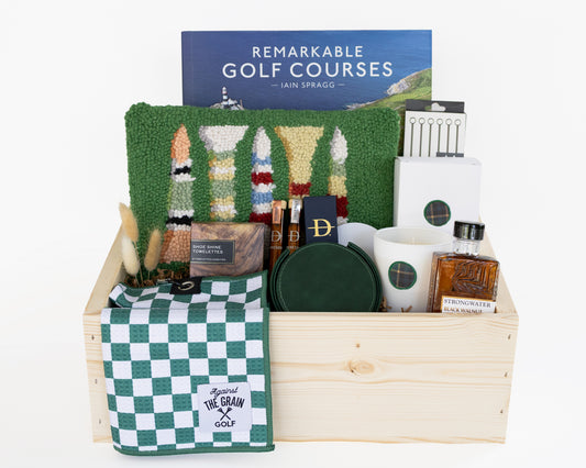 A curated gift set of golf-themed gifts on a white background, including a book titled 'Remarkable Golf Courses' by Iain Spragg, a green textured pillow with multi-colored golf tees, a green checkered towels, a white candle with a green plaid wax seal, bourbon-soaked toothpicks in both glass jars and cardboard packaging, a shoe shine kit in a wood grain box, silver plated cocktail picks, a bottle of black walnut cocktail bitters, and a set of green leather coasters. 