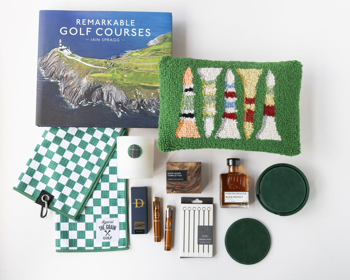 A curated flat lay of golf-themed merchandise on a white background, including a book titled 'Remarkable Golf Courses' by Iain Spragg, a green textured pillow with multi-colored golf tees, a green checkered towels, a white candle with a green plaid wax seal, bourbon-soaked toothpicks in both glass jars and cardboard packaging, a shoe shine kit in a wood grain box, silver plated cocktail picks, a bottle of black walnut cocktail bitters, and a set of green leather coasters. 