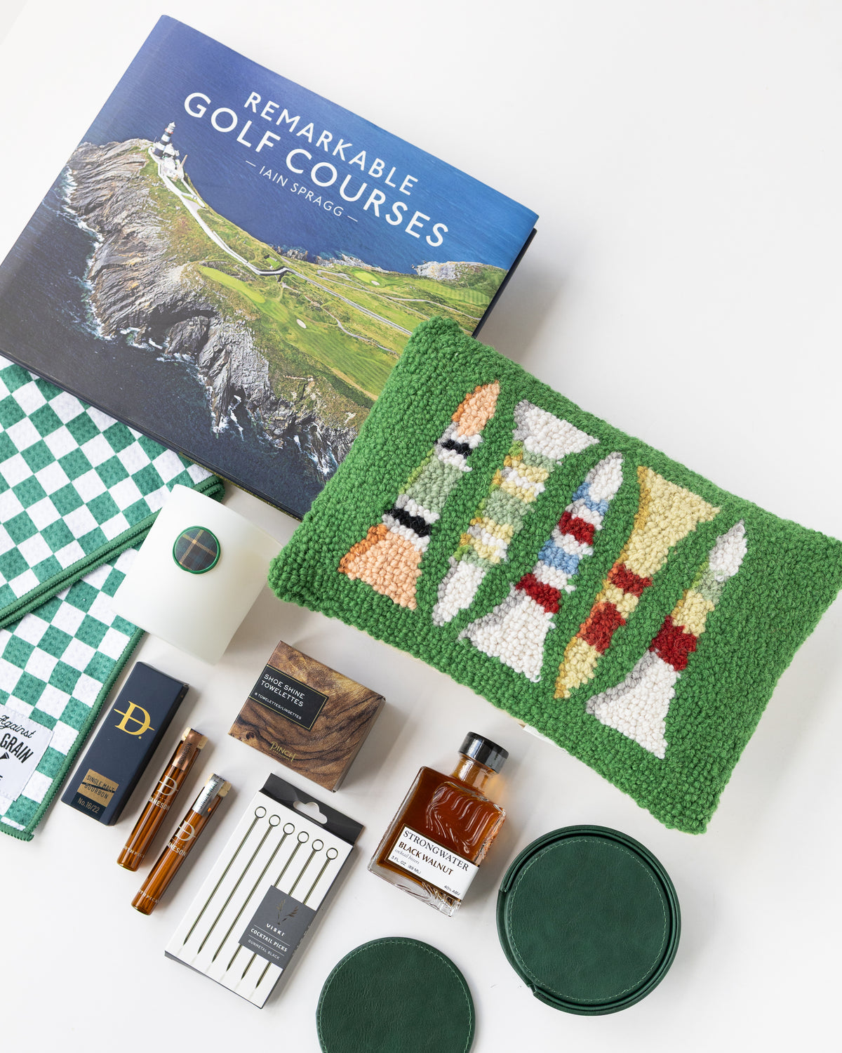 A curated flat lay of golf-themed merchandise on a white background, including a book titled 'Remarkable Golf Courses' by Iain Spragg, a green textured pillow with multi-colored golf tees, a green checkered towels, a white candle with a green plaid wax seal, bourbon-soaked toothpicks in both glass jars and cardboard packaging, a shoe shine kit in a wood grain box, silver plated cocktail picks, a bottle of black walnut cocktail bitters, and a set of green leather coasters. 