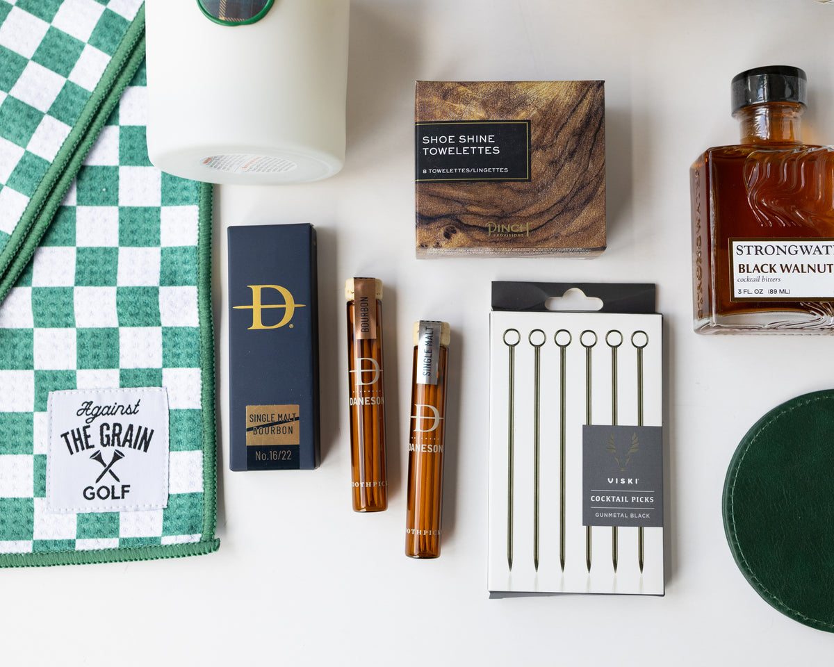 A curated flat lay of golf-themed merchandise on a white background, including a book titled 'Remarkable Golf Courses' by Iain Spragg, a green textured pillow with multi-colored golf tees, a green checkered towels, a white candle with a green plaid wax seal, bourbon-soaked toothpicks in both glass jars and cardboard packaging, a shoe shine kit in a wood grain box, silver plated cocktail picks, a bottle of black walnut cocktail bitters, and a set of green leather coasters. 