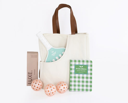 A cream canvas pickleball bag with brown leather handles, styled with a mint green and white striped pickleball paddle, three peach-orange pickleballs, a brown pickleball package labeled LUXE, and a green-and-white checkered pickleball book, all on a white background.