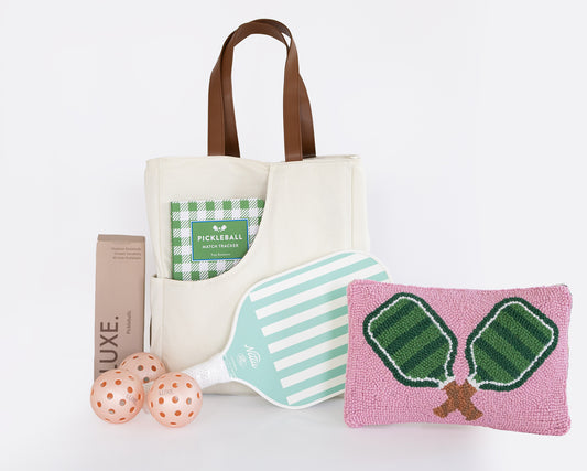 A coordinated pickleball gift set on a white background, including a cream canvas tote bag with brown handles, a green and white checkered pickleball book, a mint-and-white striped pickleball paddle, a pink woven pickleball pillow with green net embroidery, three peach-orange pickleballs, and a brown box labeled LUXE.