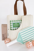 Load image into Gallery viewer, A cream canvas pickleball bag with brown leather handles, styled with a mint green and white striped pickleball paddle, three peach-orange pickleballs, a brown pickleball package labeled LUXE, and a green-and-white checkered pickleball book in its front pocket, all on a white background.

