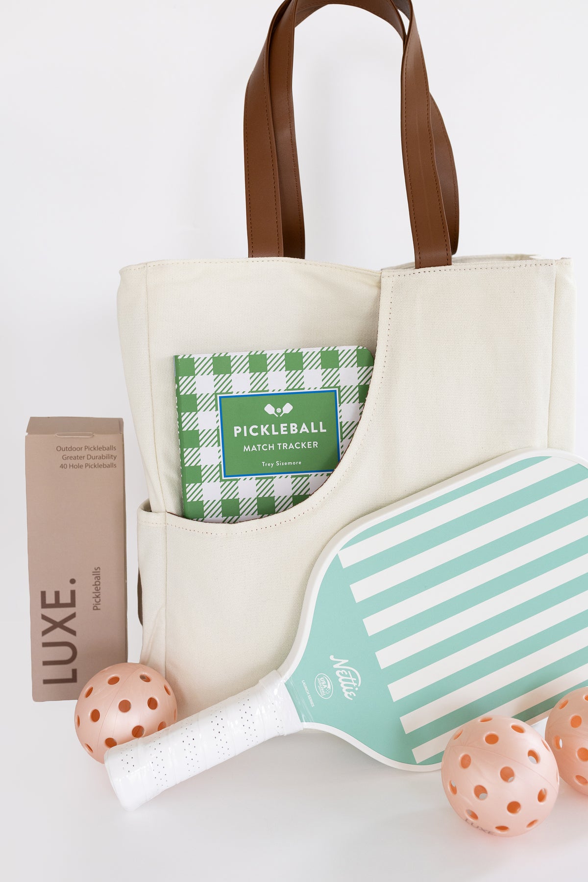 A cream canvas pickleball bag with brown leather handles, styled with a mint green and white striped pickleball paddle, three peach-orange pickleballs, a brown pickleball package labeled LUXE, and a green-and-white checkered pickleball book in its front pocket, all on a white background.