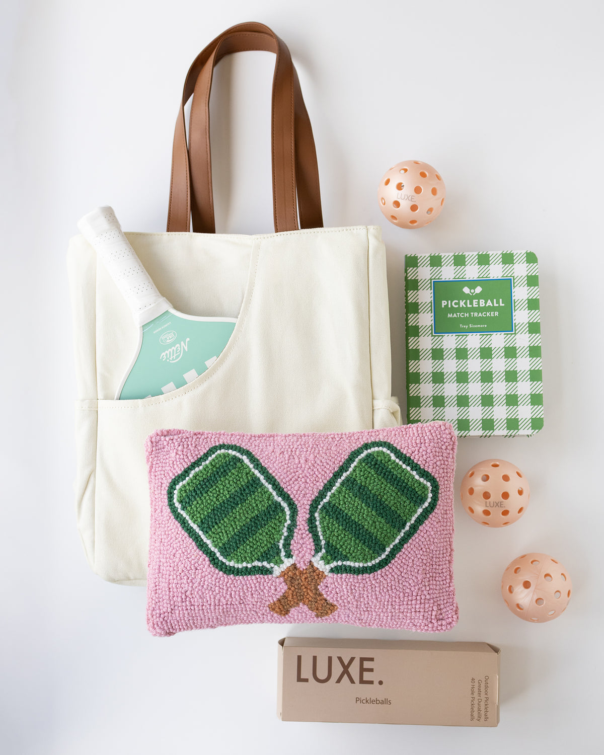 A coordinated pickleball gift set on a white background, including a cream canvas tote bag with brown handles, a green and white checkered pickleball book, a mint-and-white striped pickleball paddle, a pink woven pickleball pillow with green net embroidery, three peach-orange pickleballs, and a brown box labeled LUXE.