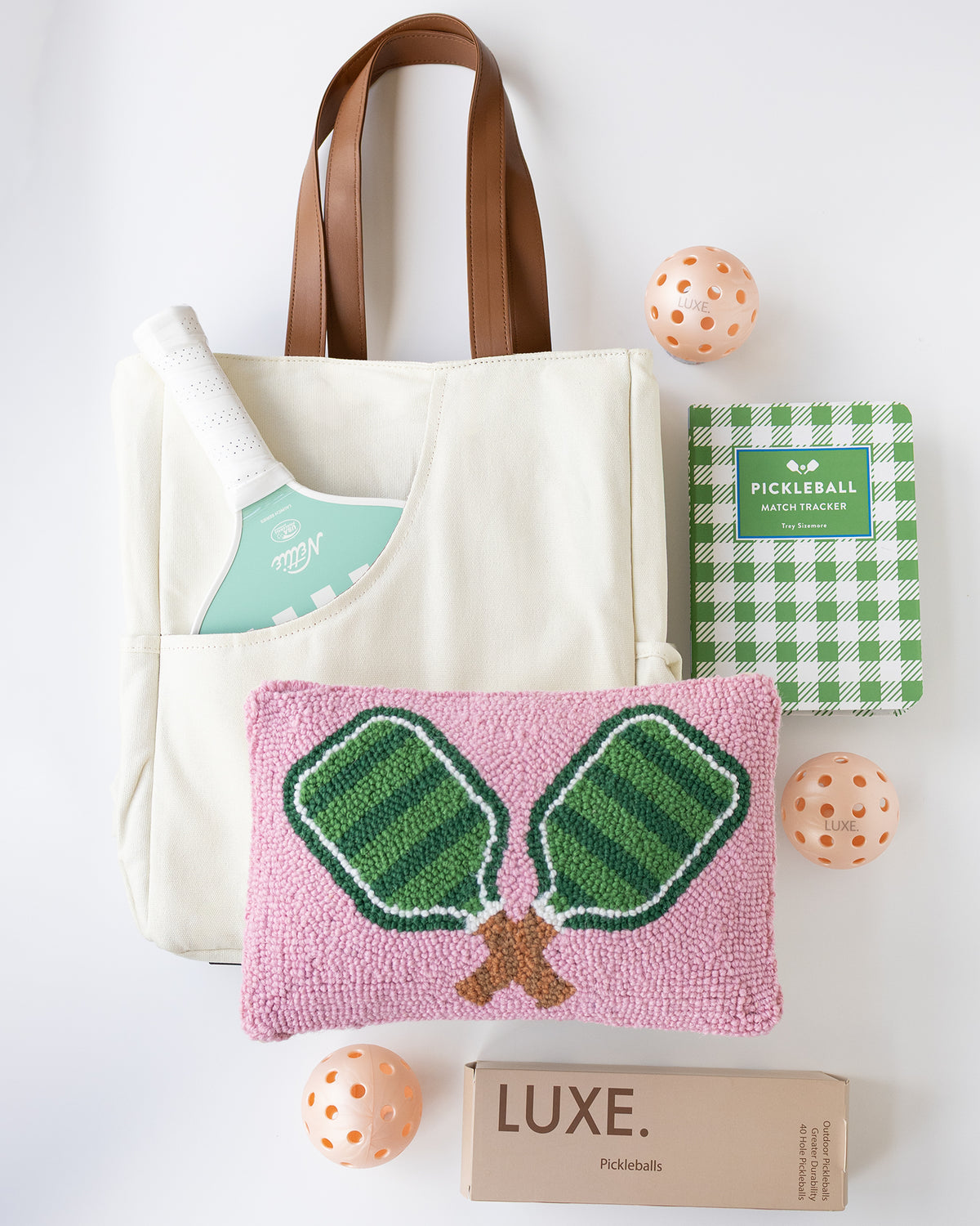 A coordinated pickleball gift set on a white background, including a cream canvas tote bag with brown handles, a green and white checkered pickleball book, a mint-and-white striped pickleball paddle, a pink woven pickleball pillow with green net embroidery, three peach-orange pickleballs, and a brown box labeled LUXE.