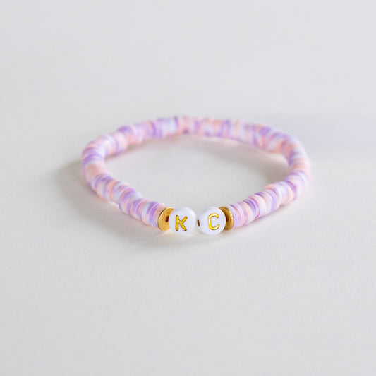 A beaded bracelet with pastel pink, purple, and white marbled beads, featuring two white round beads with gold lettering 'K' and 'C' separated by small gold spacer beads, on a light gray background.