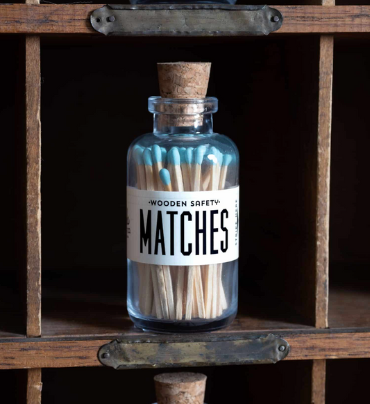 A clear glass bottle with a cork stopper filled with wooden safety matches, labeled 'WOODEN SAFETY MATCHES', displayed on a rustic wooden shelf.
